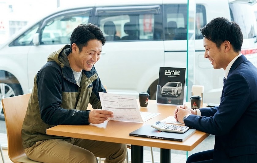 三菱ディーラーでデリカD:5の見積書を見て、笑顔で商談する男性と営業マン